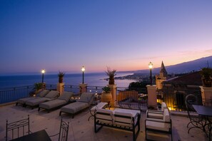 A terrace with lounge chairs and tables, overlooking a cityscape and the sea at dusk. - Casa Turchetti - Taormina (Taormina)