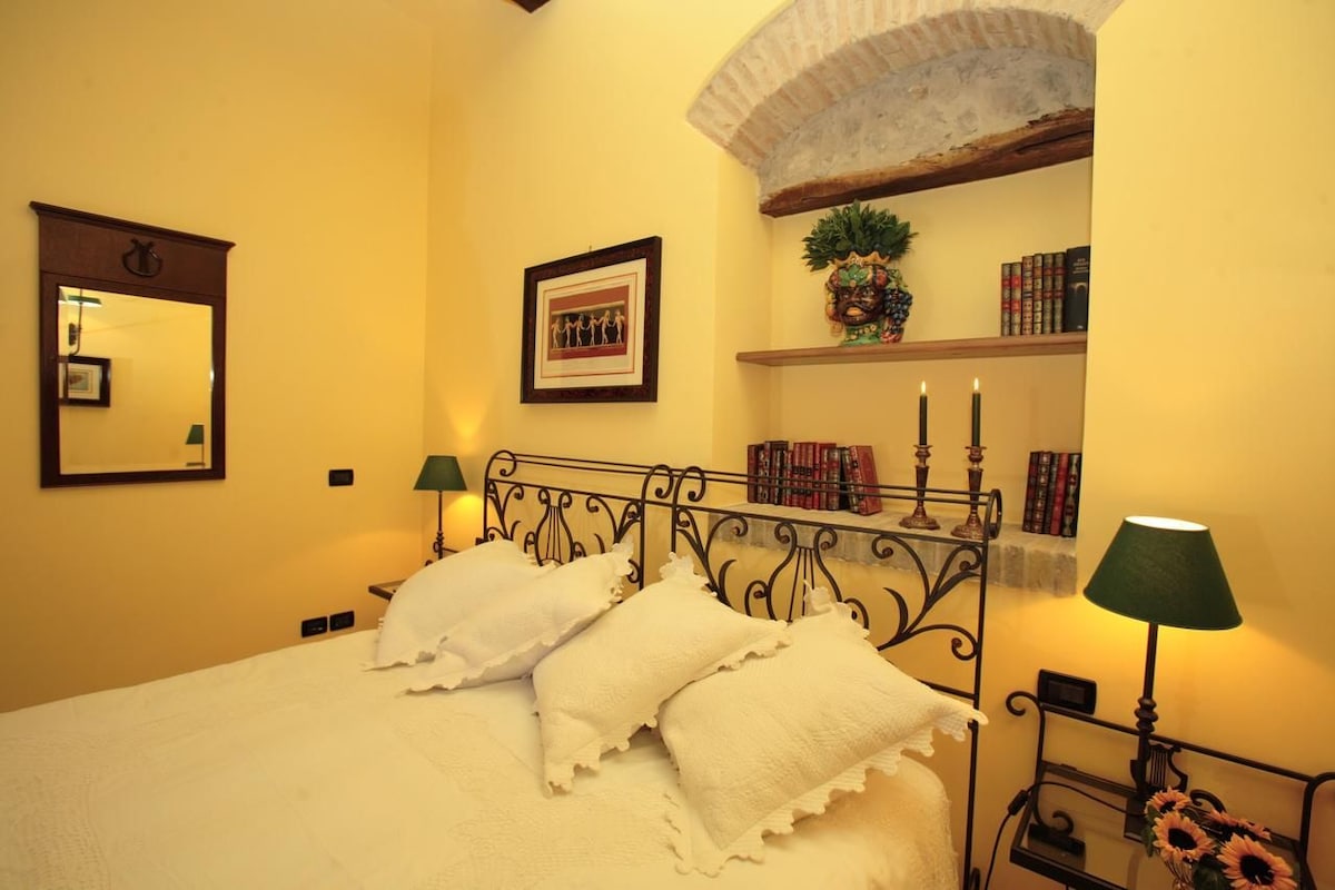 A neatly made bed with white linens and pillows, a metal headboard, a wall-mounted mirror, a framed picture, a shelf with books and a plant, and two bedside lamps.