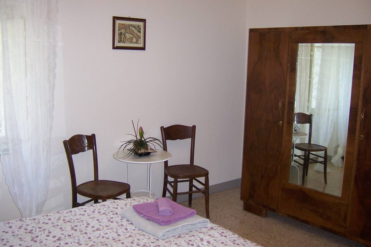 Een slaapkamer met een bed, twee stoelen, een kleine tafel met een plant, een houten deur en een lijst met een foto aan de muur.