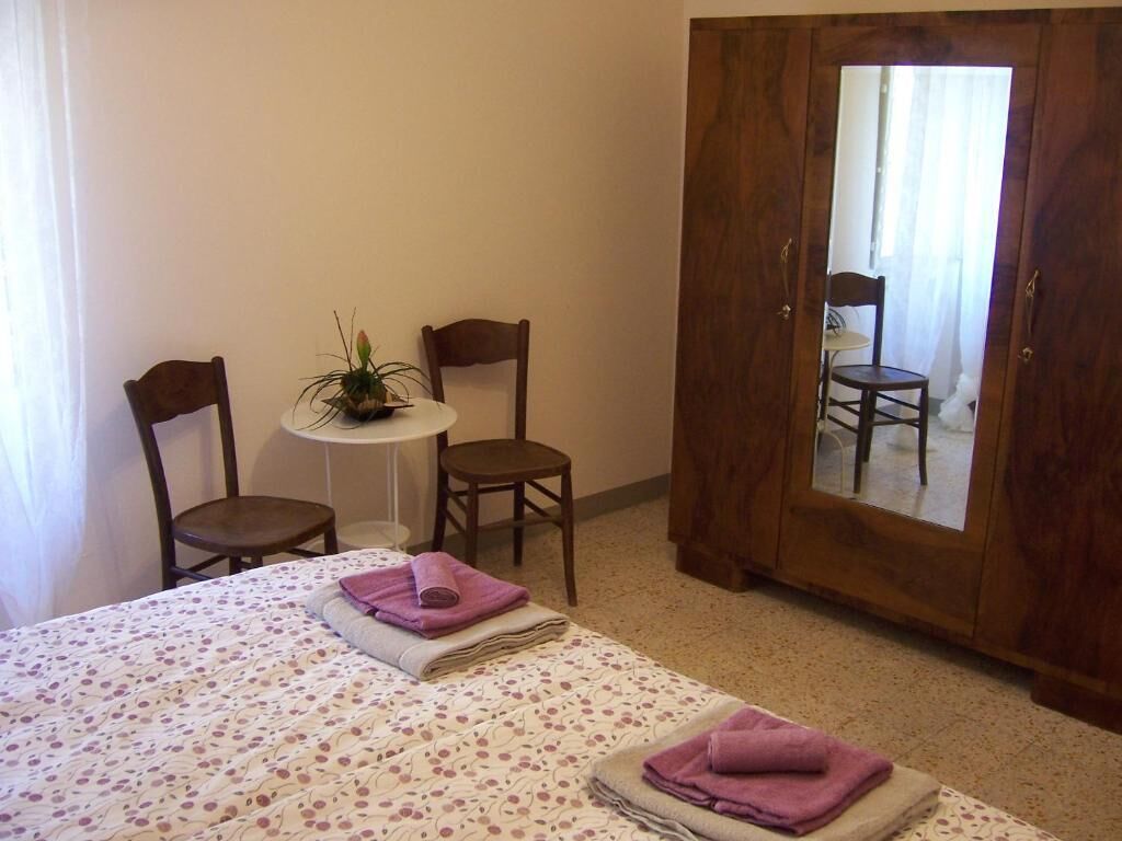 Een slaapkamer met een bed, twee stoelen, een kleine tafel met een plant, en een spiegel op een houten kast.