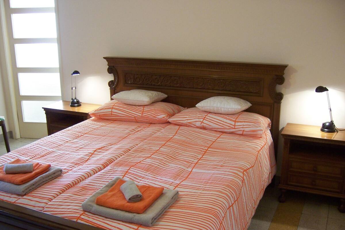 Een opgemaakt bed met gestreept beddengoed, twee nachtkastjes met lampen en een raam met zonwering.