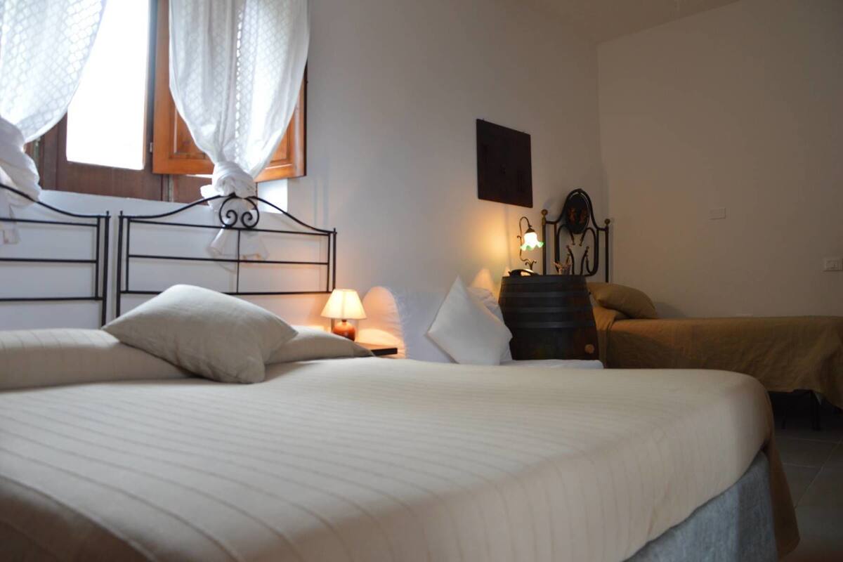 A bedroom with a large bed, a smaller bed, a nightstand with a lamp, a wall-mounted shelf, and a window with a curtain.