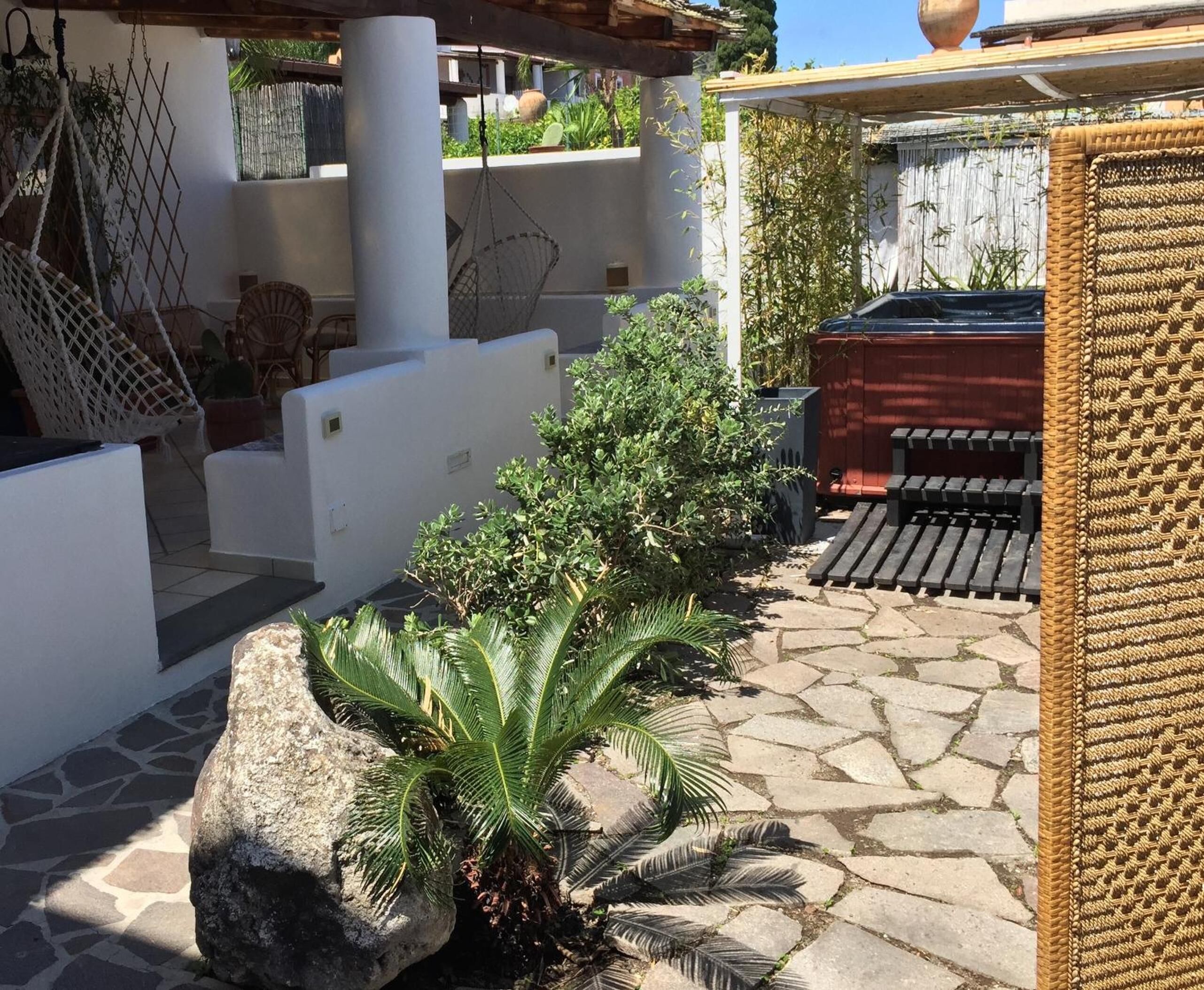 Un patio con suelo de piedra, una maceta de roca grande con una planta de palma, una silla colgante y un banco tejido.