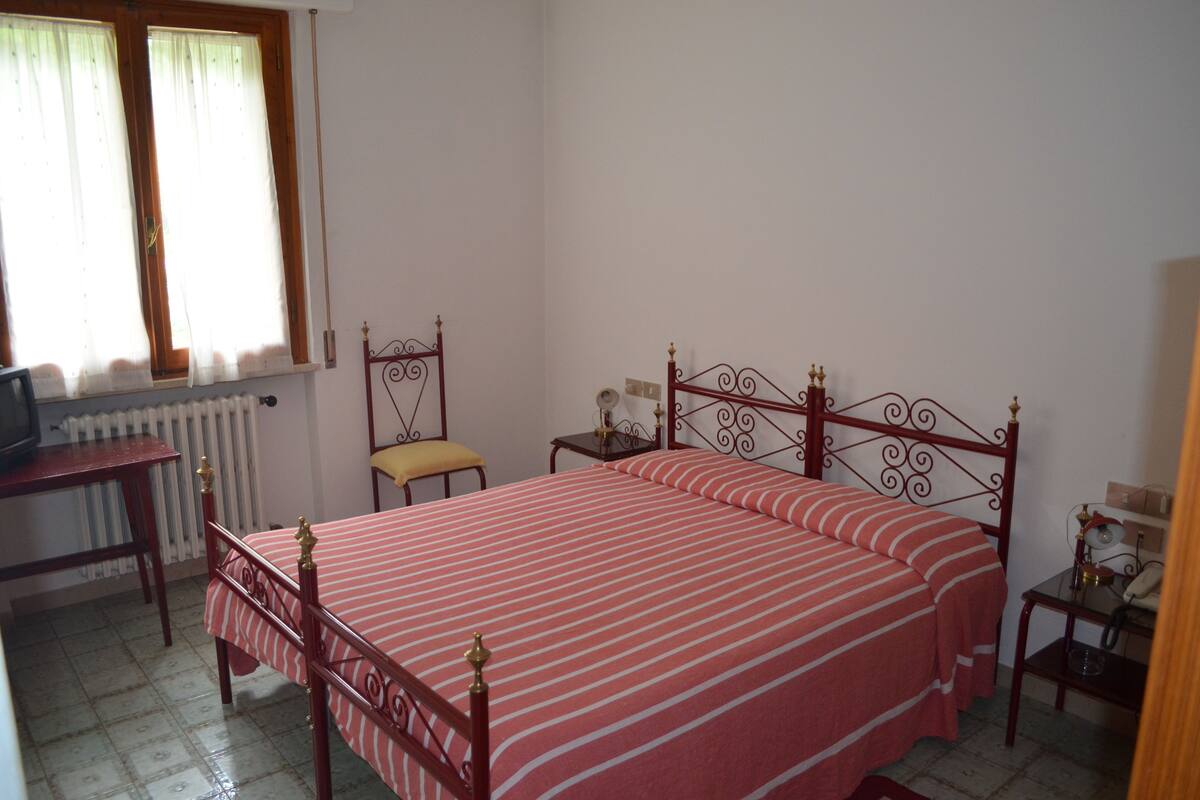 A bedroom with a bed, a chair, a small table, a radiator, and a window with curtains.