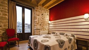 Un dormitorio con pared de piedra, pared de acento roja, cama con colcha estampada, una silla roja y vistas a través de una ventana.
