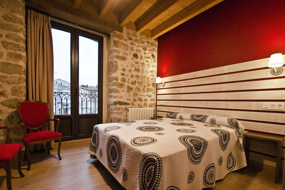 Een slaapkamer met een stenen muur, een rode accentmuur, een bed met een geborduurde spreideken, een rode stoel en een uitzicht door een raam.