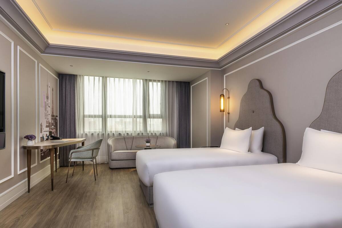 Ein Hotelzimmer mit zwei Betten, einem Schreibtisch, einem Stuhl und einem großen Fenster mit Vorhängen.