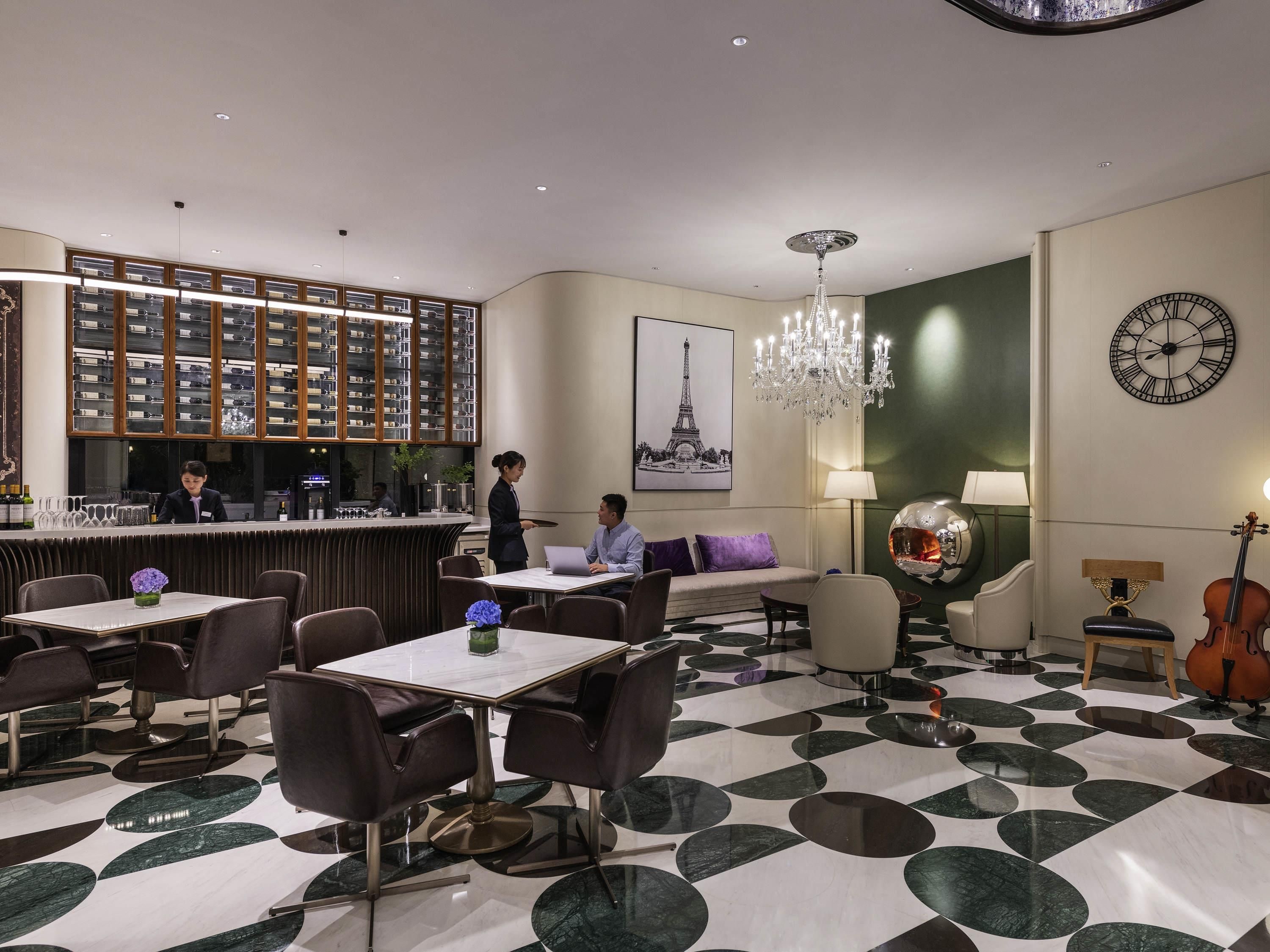 A hotel lobby with a bar area, seating arrangements, a chandelier, and a clock on the wall.