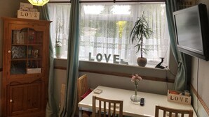 A cozy room with a wooden cabinet, a table with chairs, a TV, and a window with 'LOVE' decorations. - Love & Peace (Orpington)