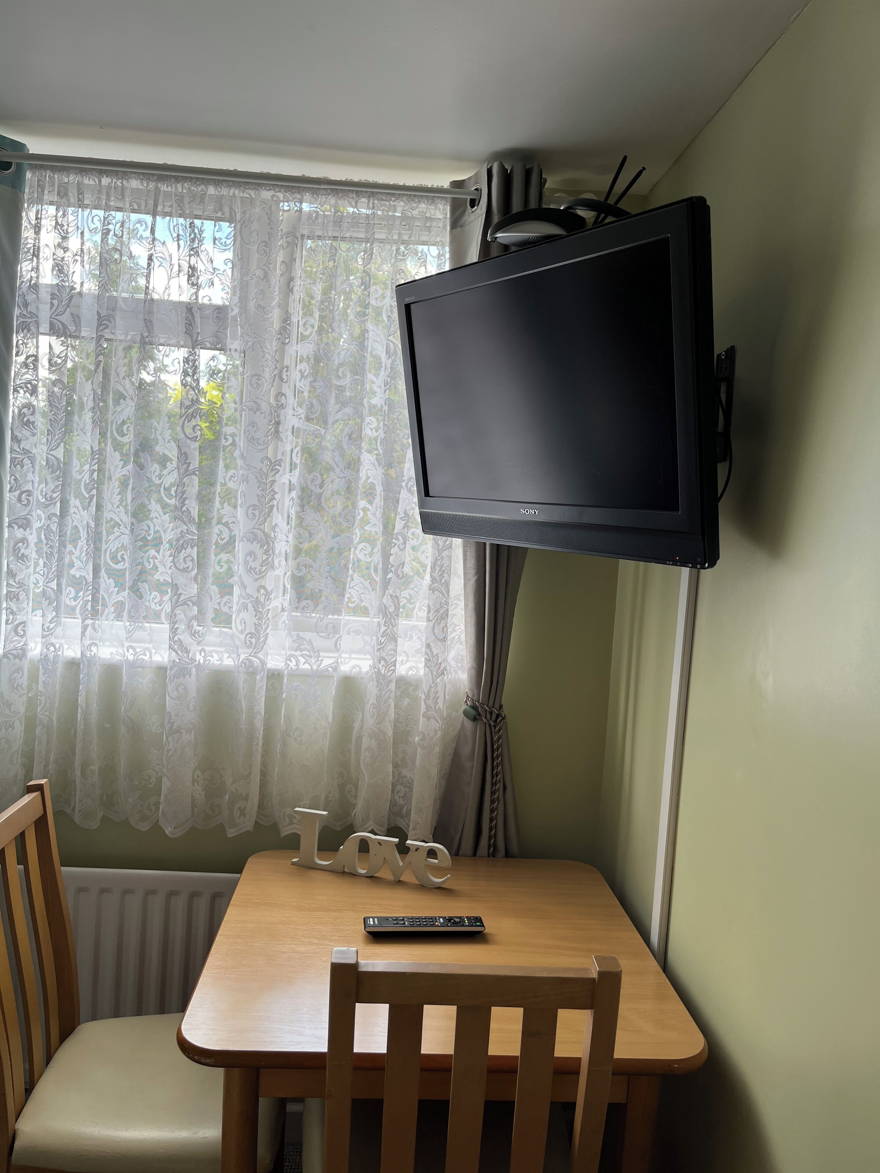 Une pièce avec une table en bois, deux chaises, un téléviseur fixé au mur et une fenêtre ornée de rideaux à motifs.