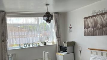A hotel room with a bed, a desk, a chair, a radiator, a microwave, a nightstand, a lamp, a water bottle, and a window with curtains.