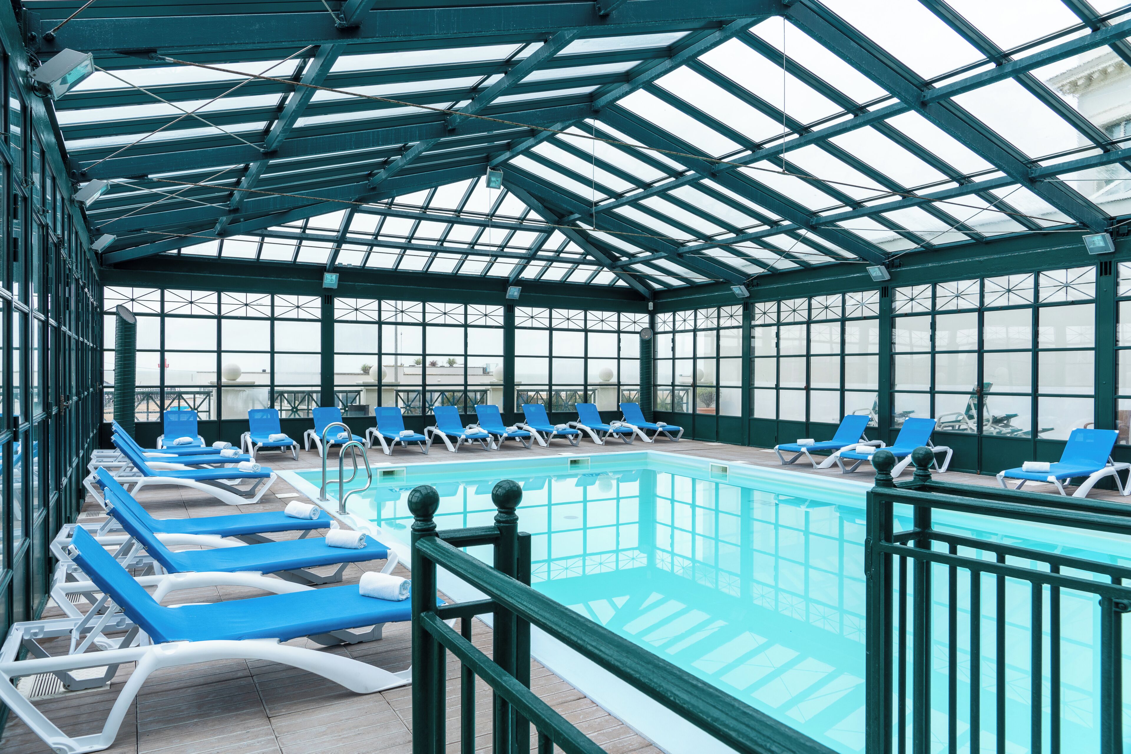 A covered swimming pool with blue lounge chairs and a glass ceiling.