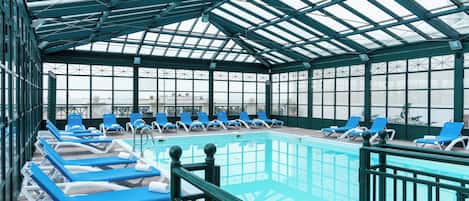 Piscina techada con sillas reclinables azules y techo de vidrio.
