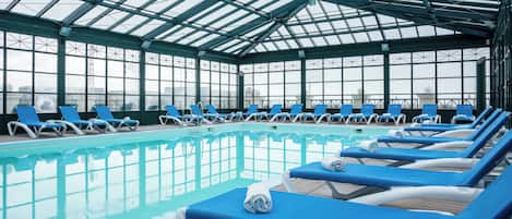 An indoor swimming pool with blue lounge chairs and a glass ceiling.