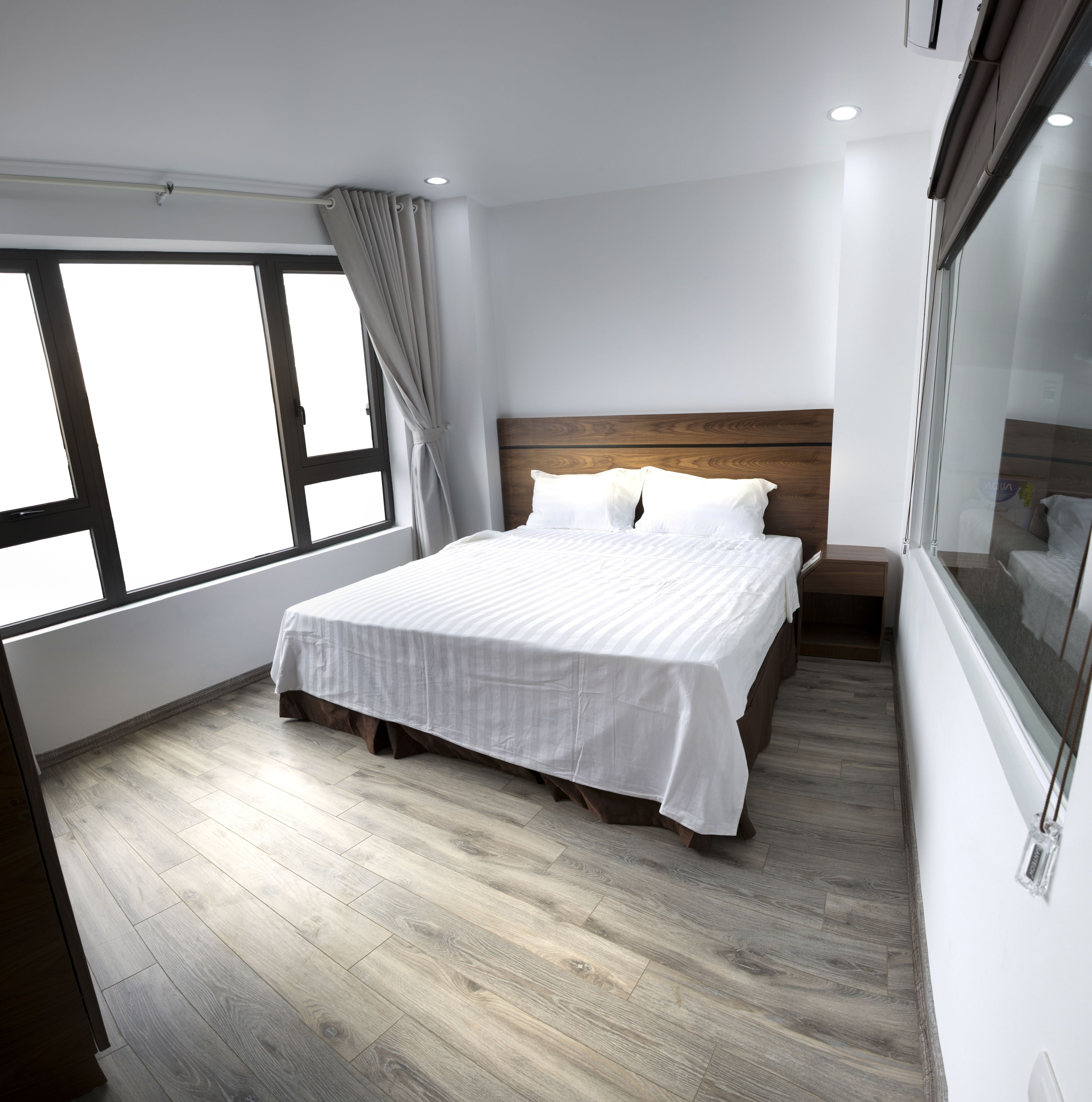 A modern bedroom with a large bed, a wooden headboard, a window with curtains, and a wooden floor.