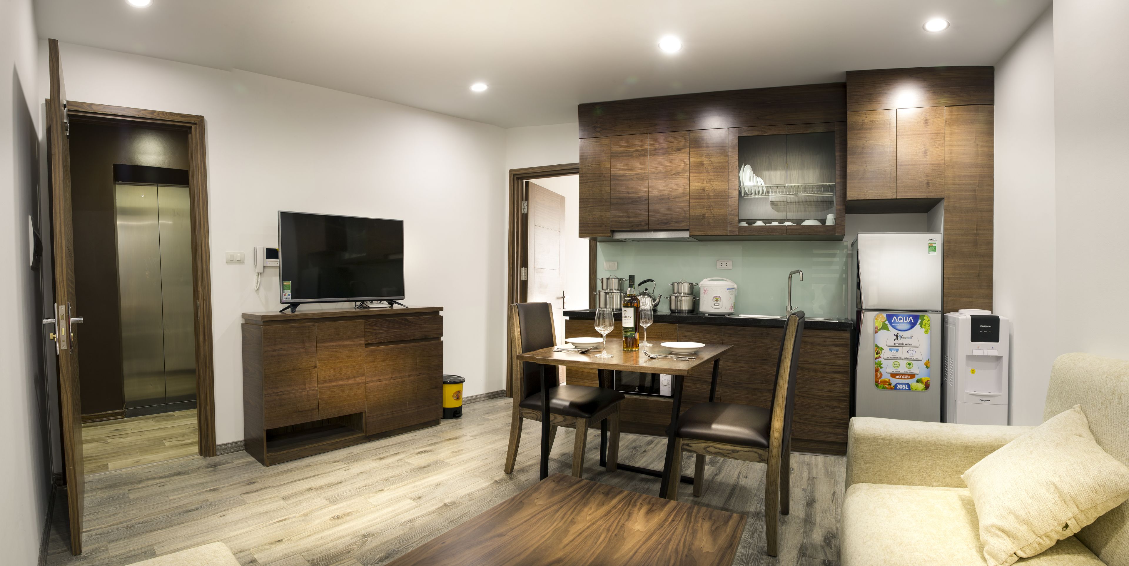 Een kleine keuken met houten kastjes, een eettafel met stoelen, een televisie en een koelkast.