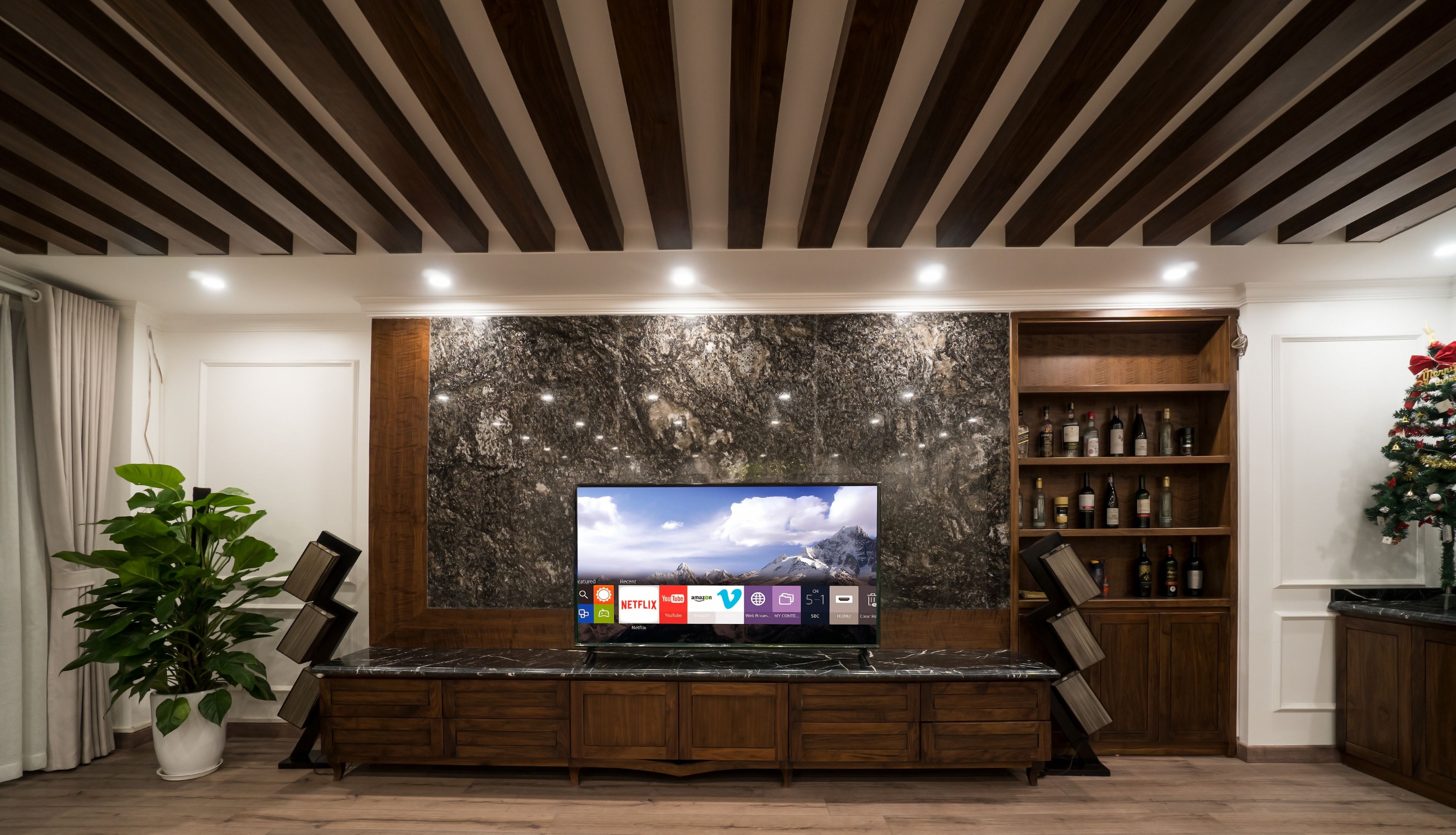 A modern living room with a large flat-screen TV mounted on a marble wall, wooden shelving with various items, and a potted plant.