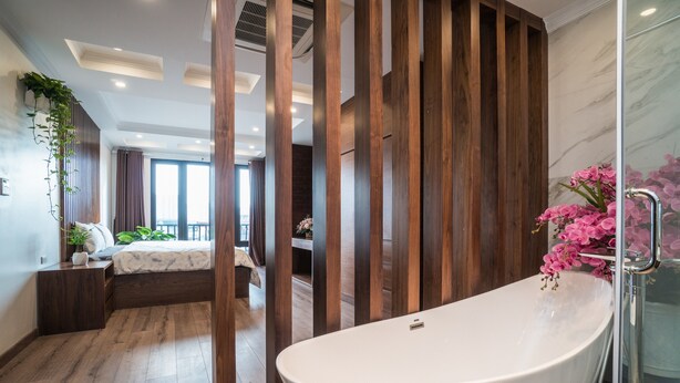 A modern bedroom with a large bed, a wooden partition, and a bathroom with a bathtub and pink flowers. - iRest Apartment (Hanoi)