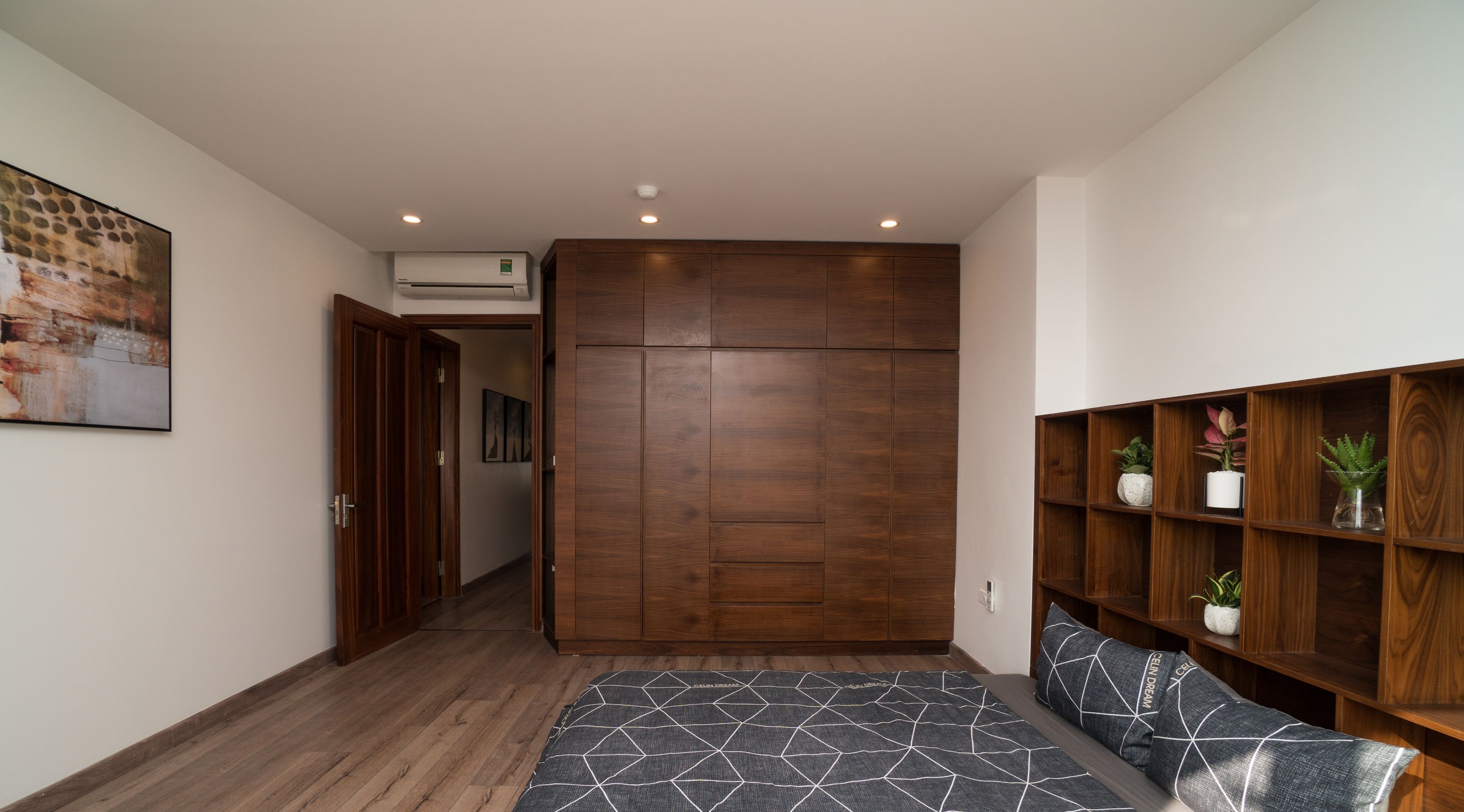 Quarto com guarda-roupa de madeira, cama com roupa de cama estampada, prateleira de madeira com plantas e um quadro na parede.