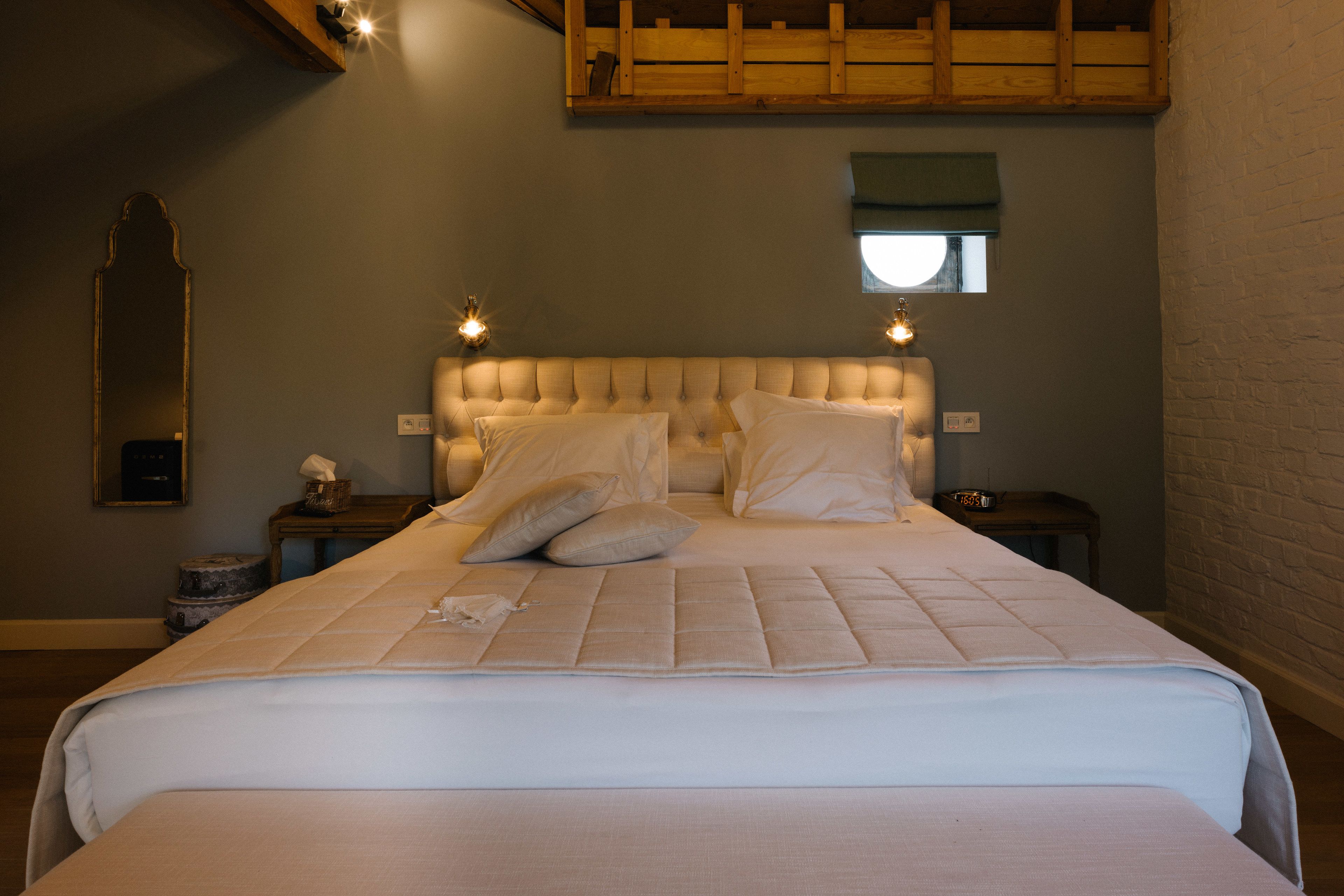 A neatly made bed with a tufted headboard, a bedside table with a lamp, and a small window with a view of the outside.