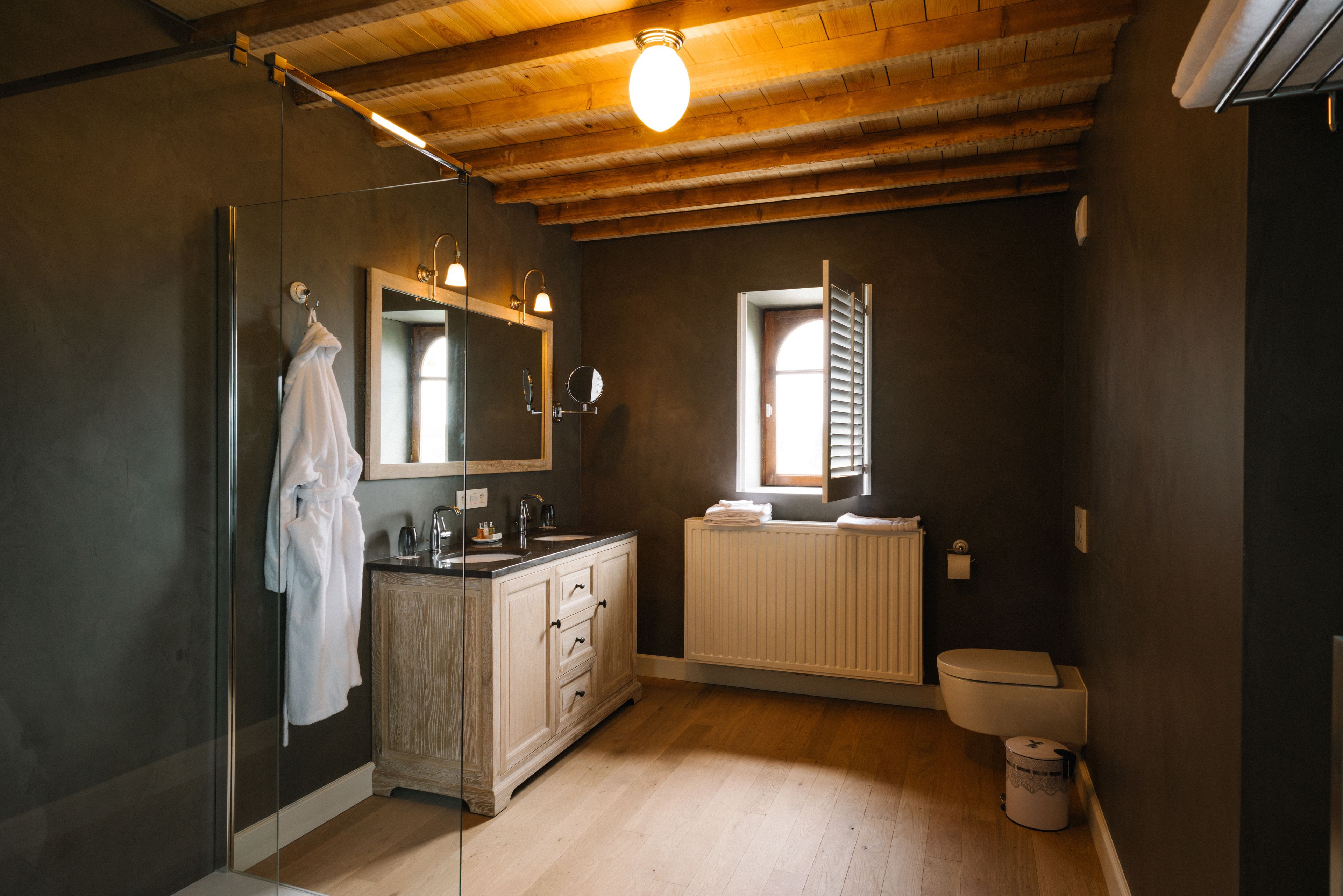 A bathroom with a glass shower enclosure, a wooden vanity with a mirror, a toilet, and a window with blinds.