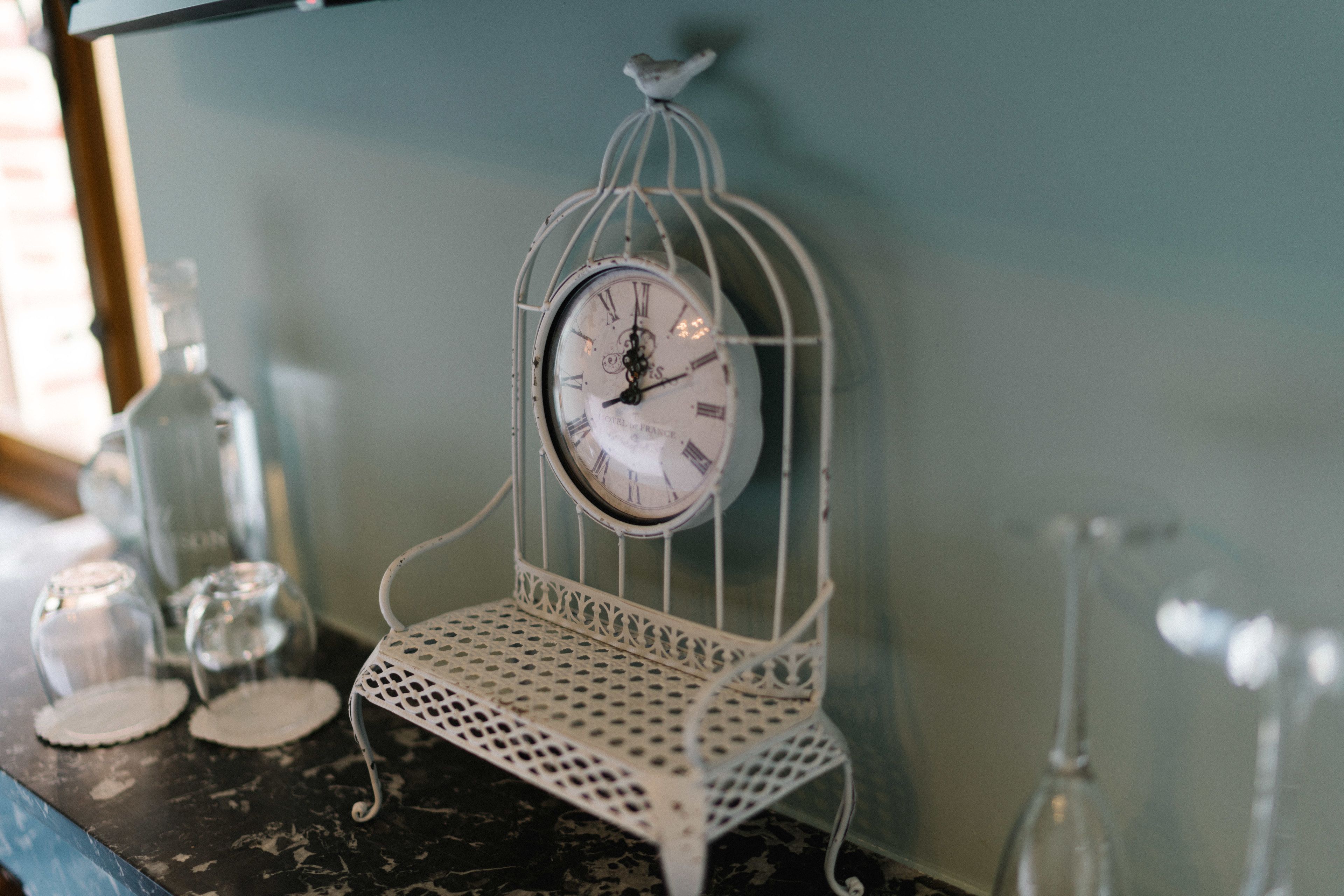 A white birdcage clock with Roman numerals on a dark surface, surrounded by clear glass bottles and a metal tray.