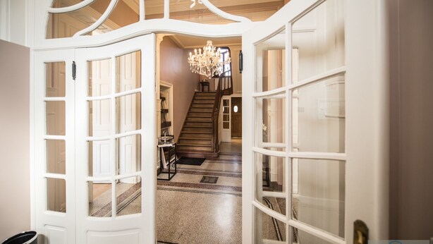 Una puerta abierta que da a una escalera con candelabro, peldaños alfombrados y vista a una habitación con una puerta y un bote de basura.