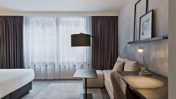 A hotel room with a bed, a sofa, a desk, a lamp, and a large window with curtains.