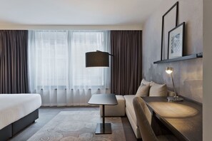 A hotel room with a bed, a sofa, a desk, a lamp, and a large window with curtains. - Residence Inn by Marriott Frankfurt City Center (Frankfurt)