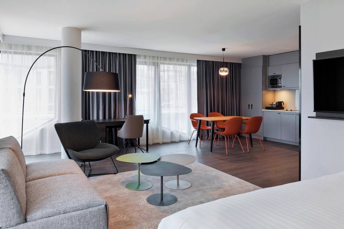 Ein modernes Hotelzimmer mit Sofa, einem Esstisch mit Stühlen, einer kleinen Küchenzeile und einem großen Fenster mit Vorhängen.