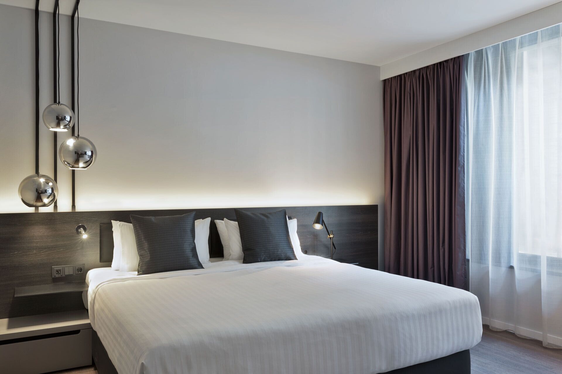 A modern hotel room with a large bed, bedside lamps, a nightstand, and a window with curtains.
