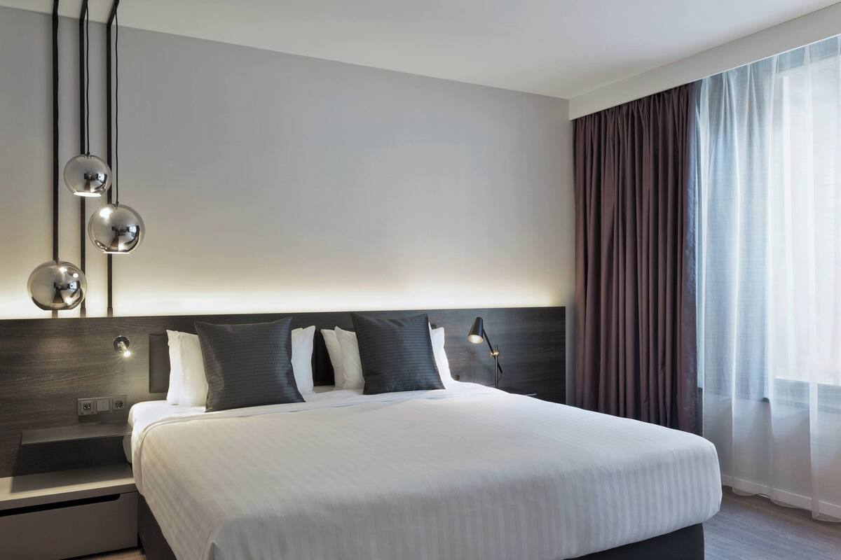 Ein modernes Hotelzimmer mit einem großen Bett, einem Nachttisch, einer Wandlampe und einem Fenster mit Vorhängen.