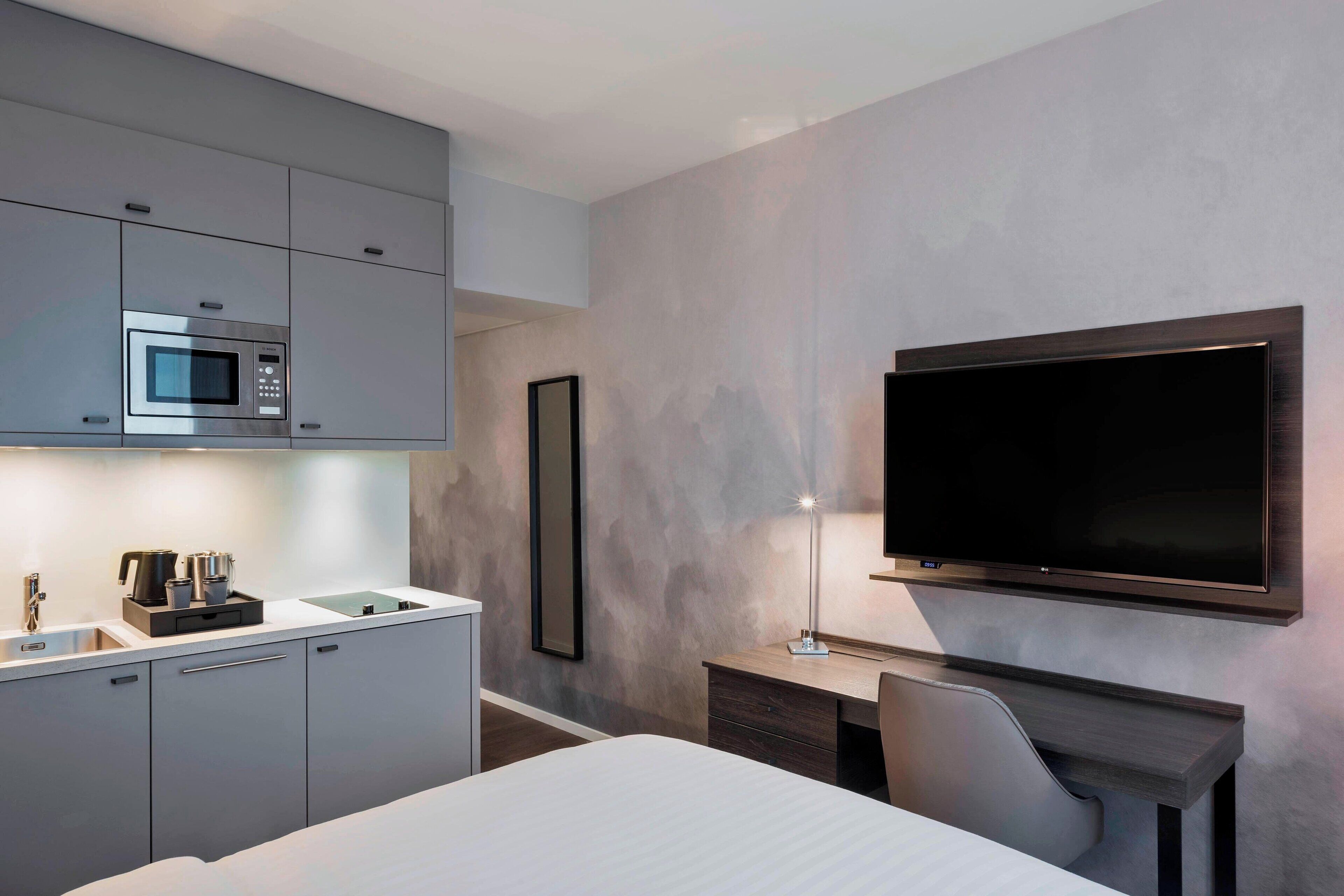 A modern hotel room with a bed, a desk with a chair, a flat-screen TV mounted on the wall, a built-in microwave, and a coffee maker.