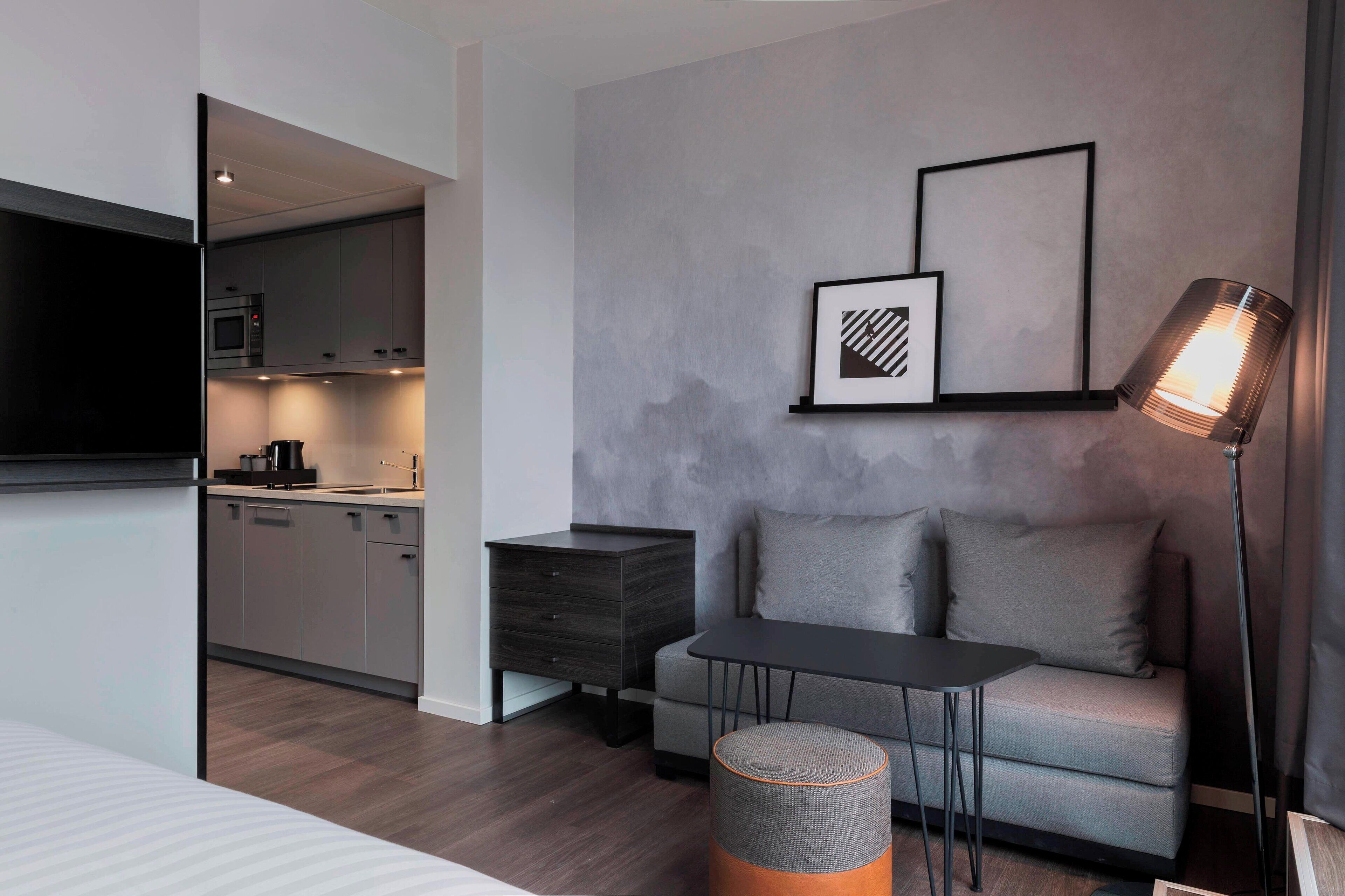 Quarto de hotel moderno com um sofá cinzento, uma mesinha, uma mesinha de cabeceira, um abajur e uma televisão.