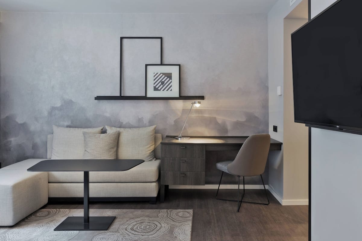 Ein modernes Wohnzimmer mit einer Couch, einem kleinen Tisch, einem Schreibtisch und einem Sessel. An der Wand ist ein Flachbildfernseher angebracht.