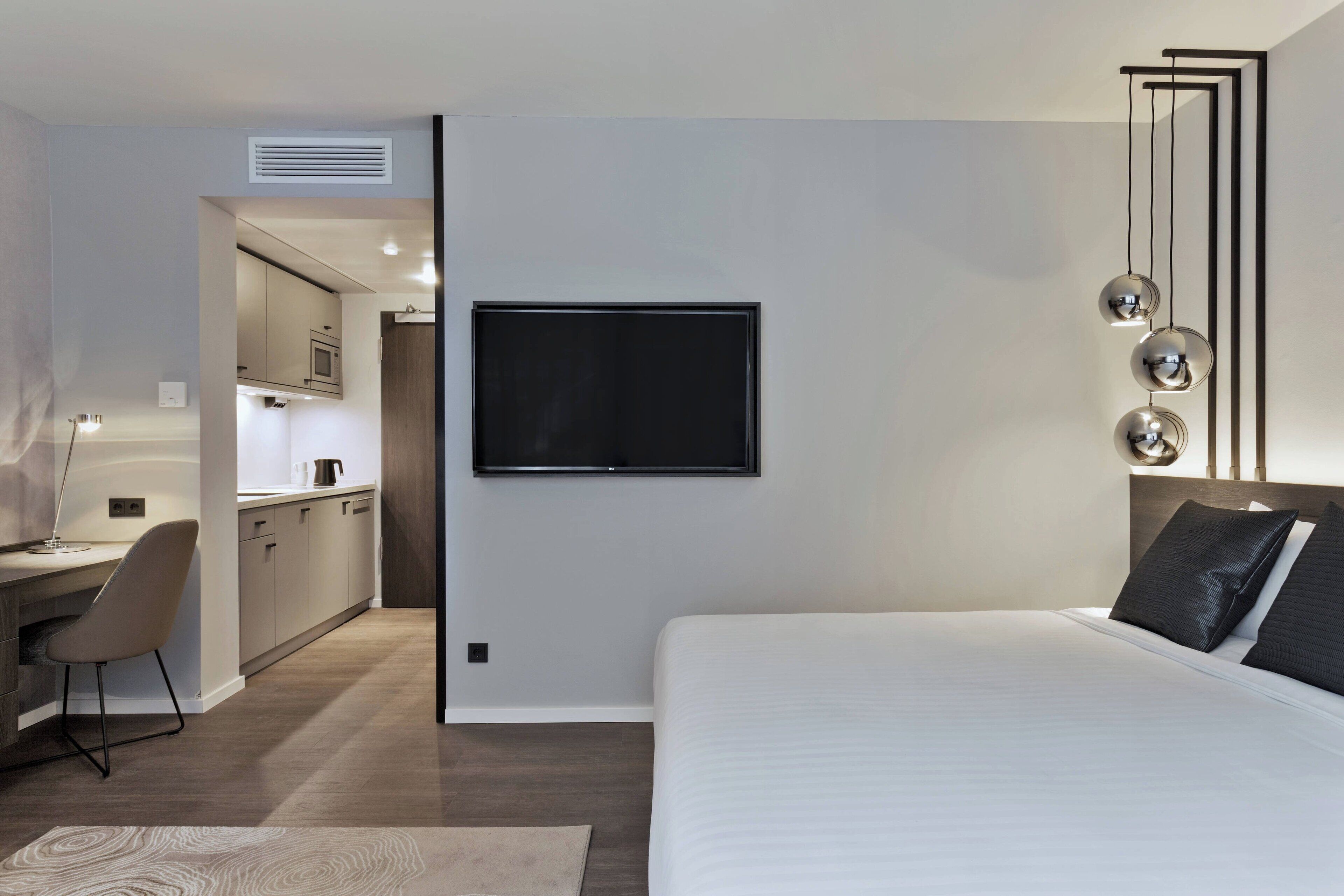 A modern hotel room with a large bed, a desk with a chair, a kitchenette, and a TV mounted on the wall.