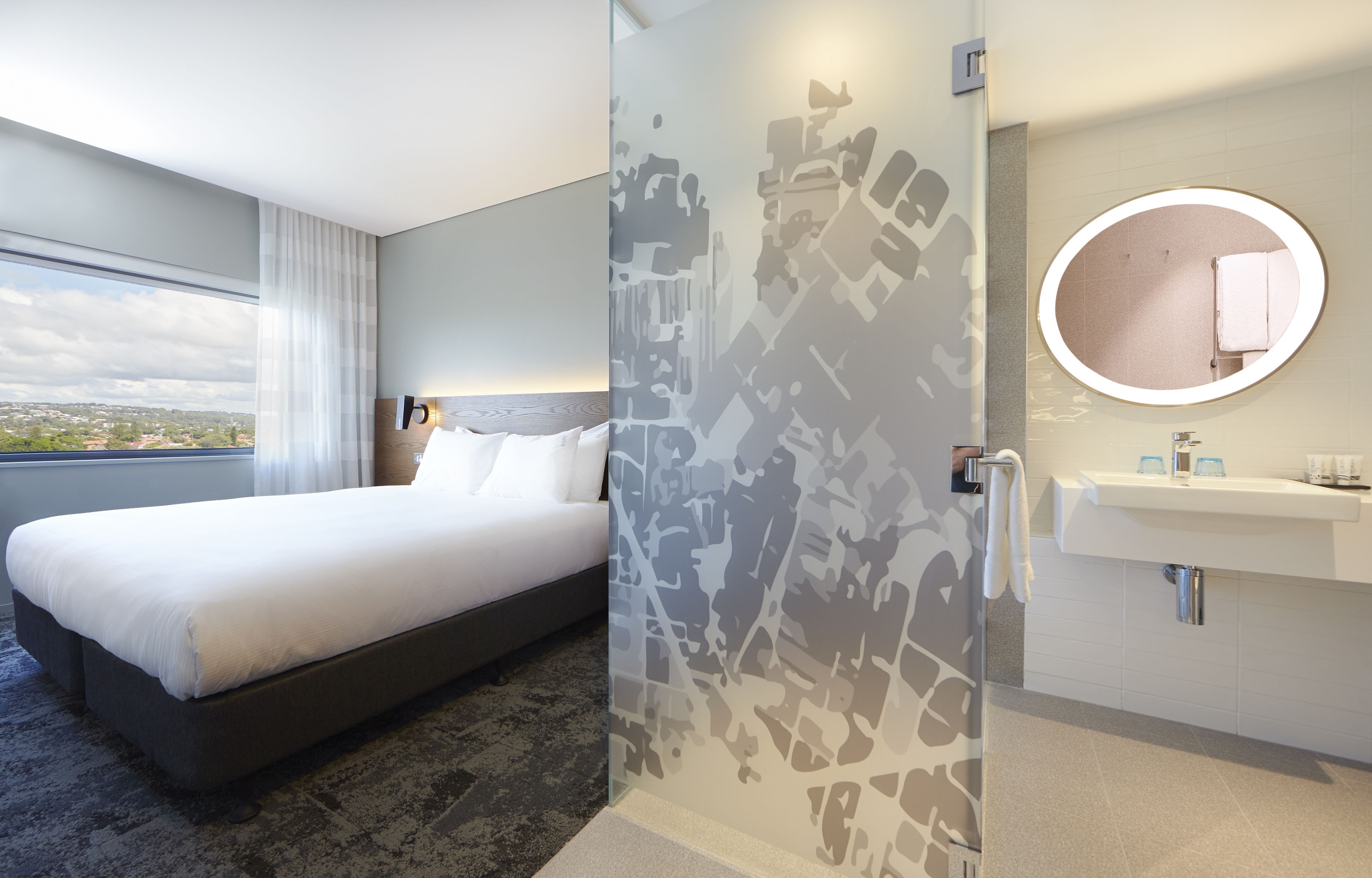 Una habitación de hotel moderna con una cama grande, cabina de ducha de vidrio, espejo redondo y vistas a la ciudad.