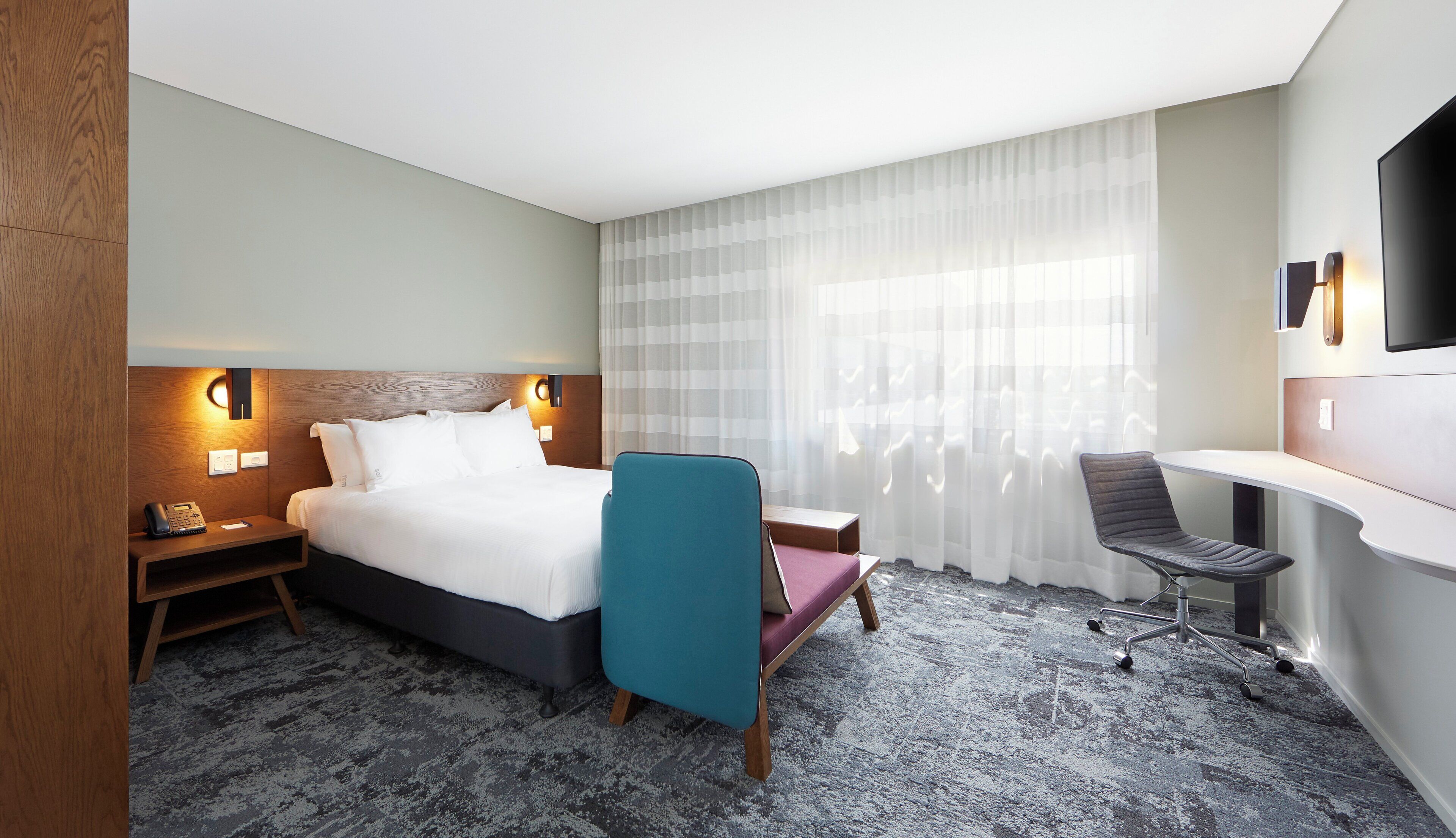 A hotel room with a bed, a desk, a chair, a telephone, and a TV.