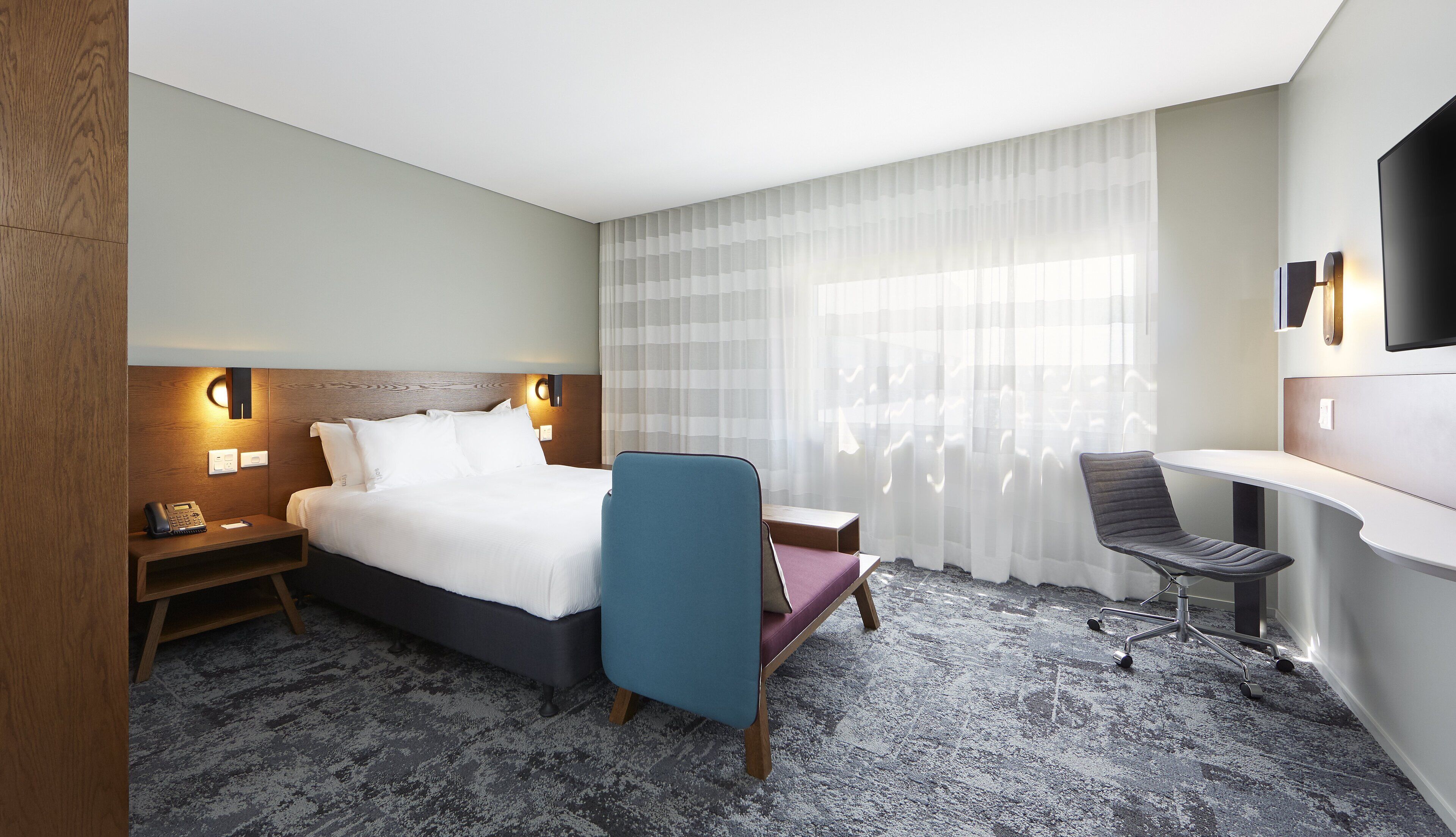 A hotel room with a bed, bedside table, lamp, chair, desk, and a TV.