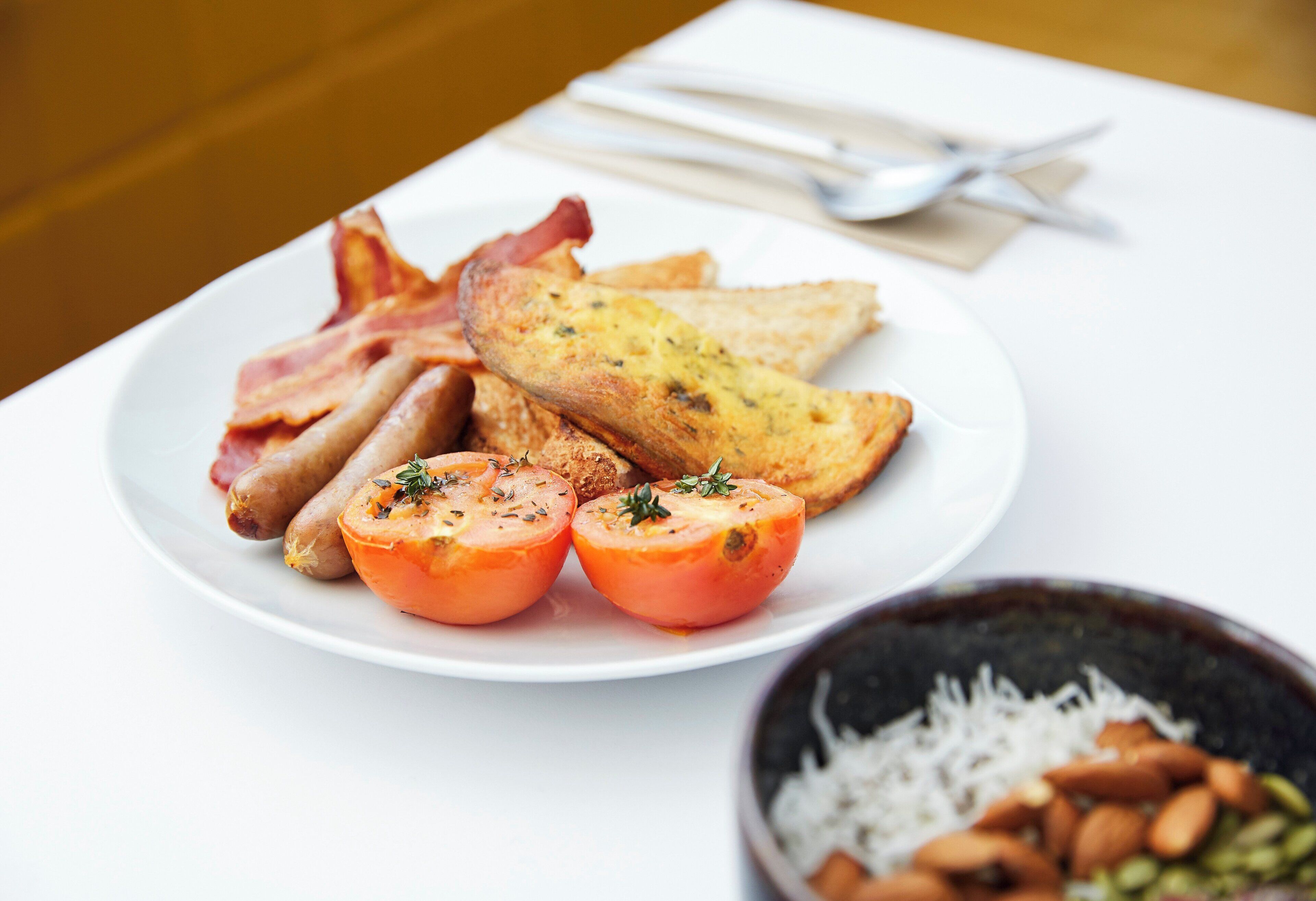 Une assiette avec des lardons, des toasts et des tomates, et un bol de riz et d’amandes sur une table.