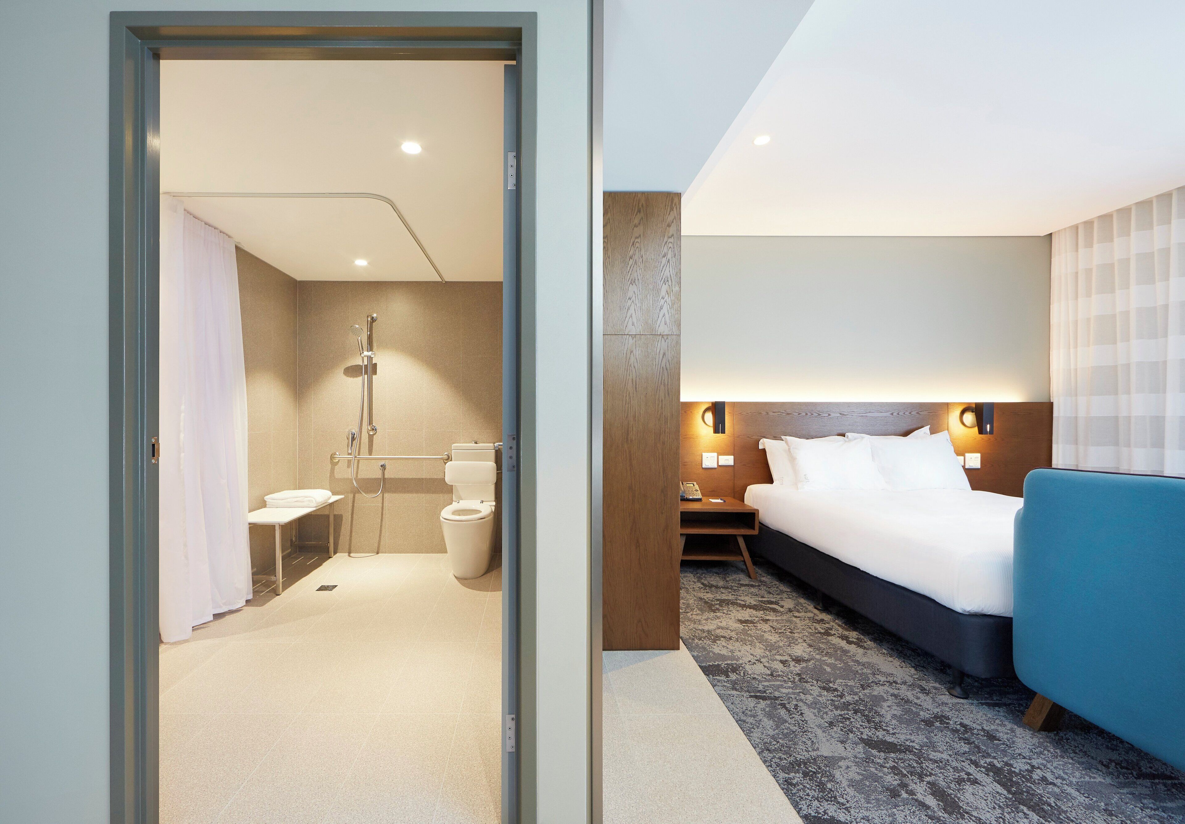 Una habitación de hotel con ducha, inodoro, cama, mesita de noche y un sillón azul.