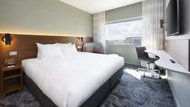 A hotel room with a large bed, a desk, a chair, a TV, and a window with a city view.