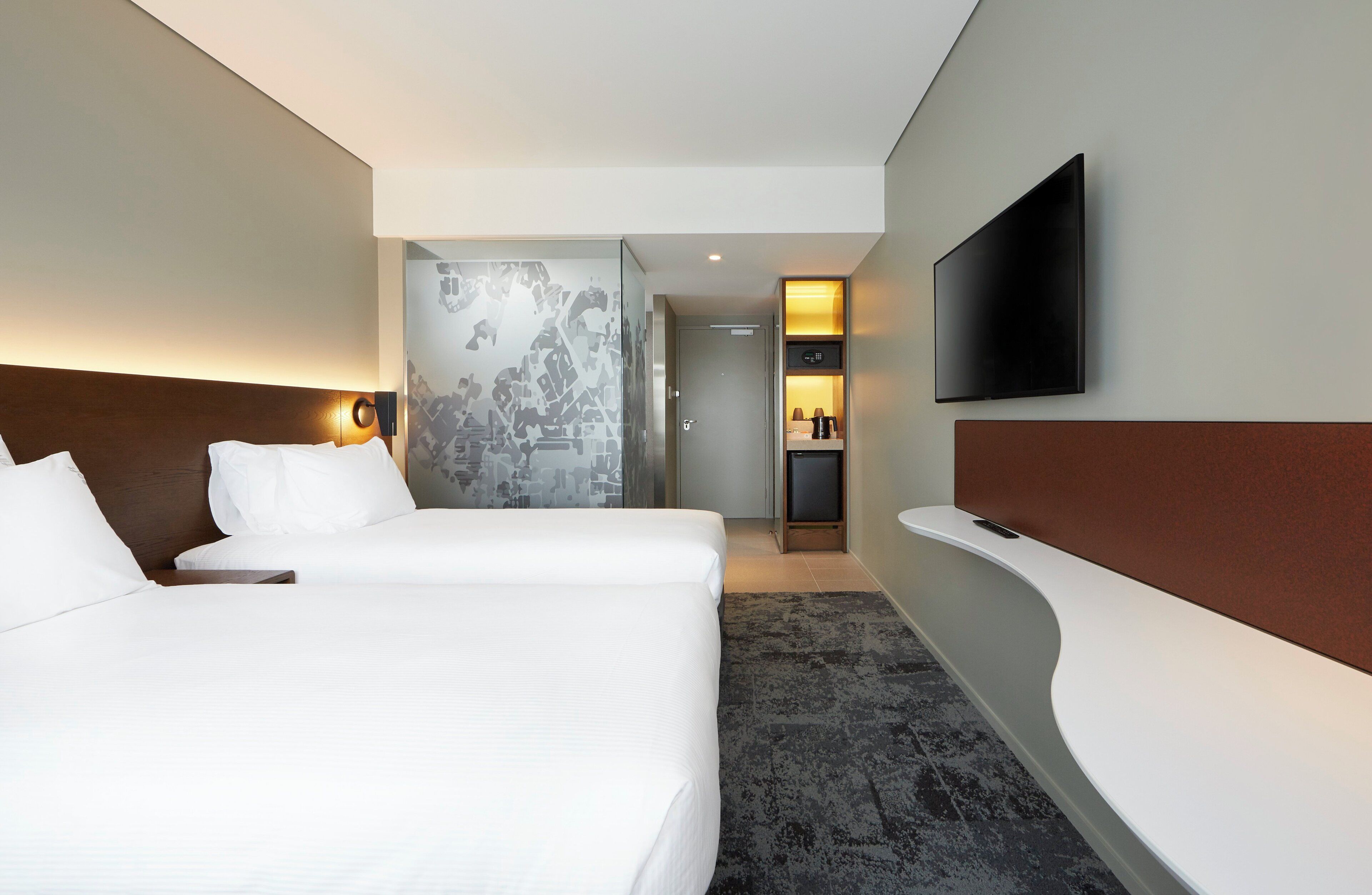 A hotel room with two beds, a flat-screen TV mounted on the wall, and a small kitchenette area.