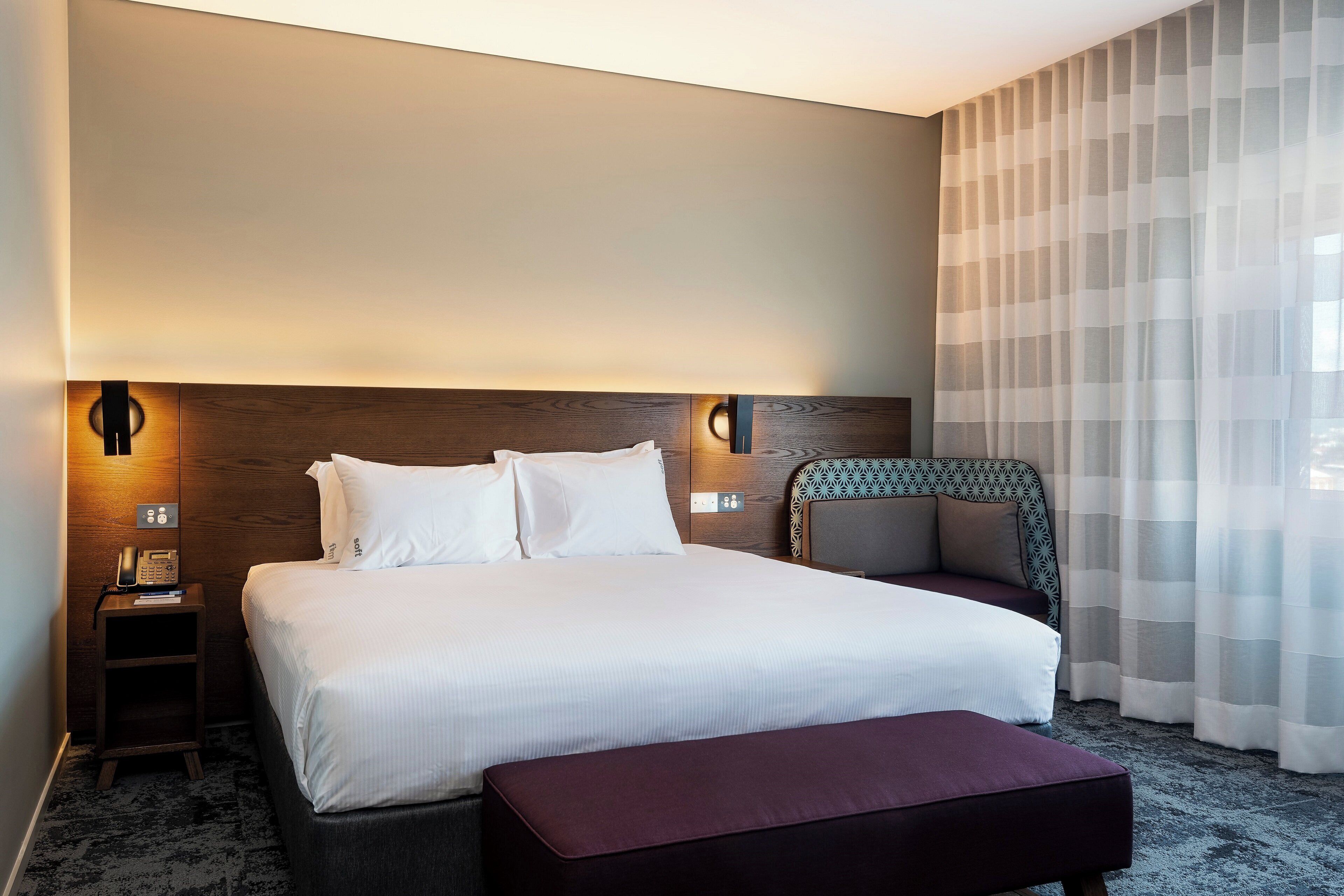 Une chambre d’hôtel avec un grand lit, des tables de chevet, une petite table, une chaise et une fenêtre avec des rideaux légers.