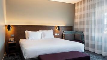 A hotel room with a large bed, bedside tables, a nightstand, a chair, and a window with sheer curtains.
