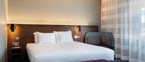A hotel room with a large bed, bedside tables, a nightstand, a chair, and a window with sheer curtains.