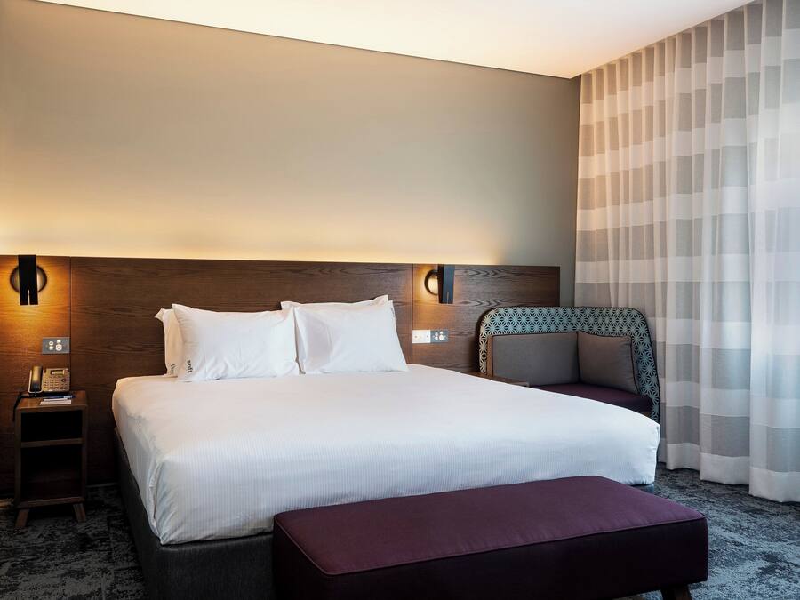 A hotel room with a large bed, bedside tables, a nightstand, a chair, and a window with sheer curtains.