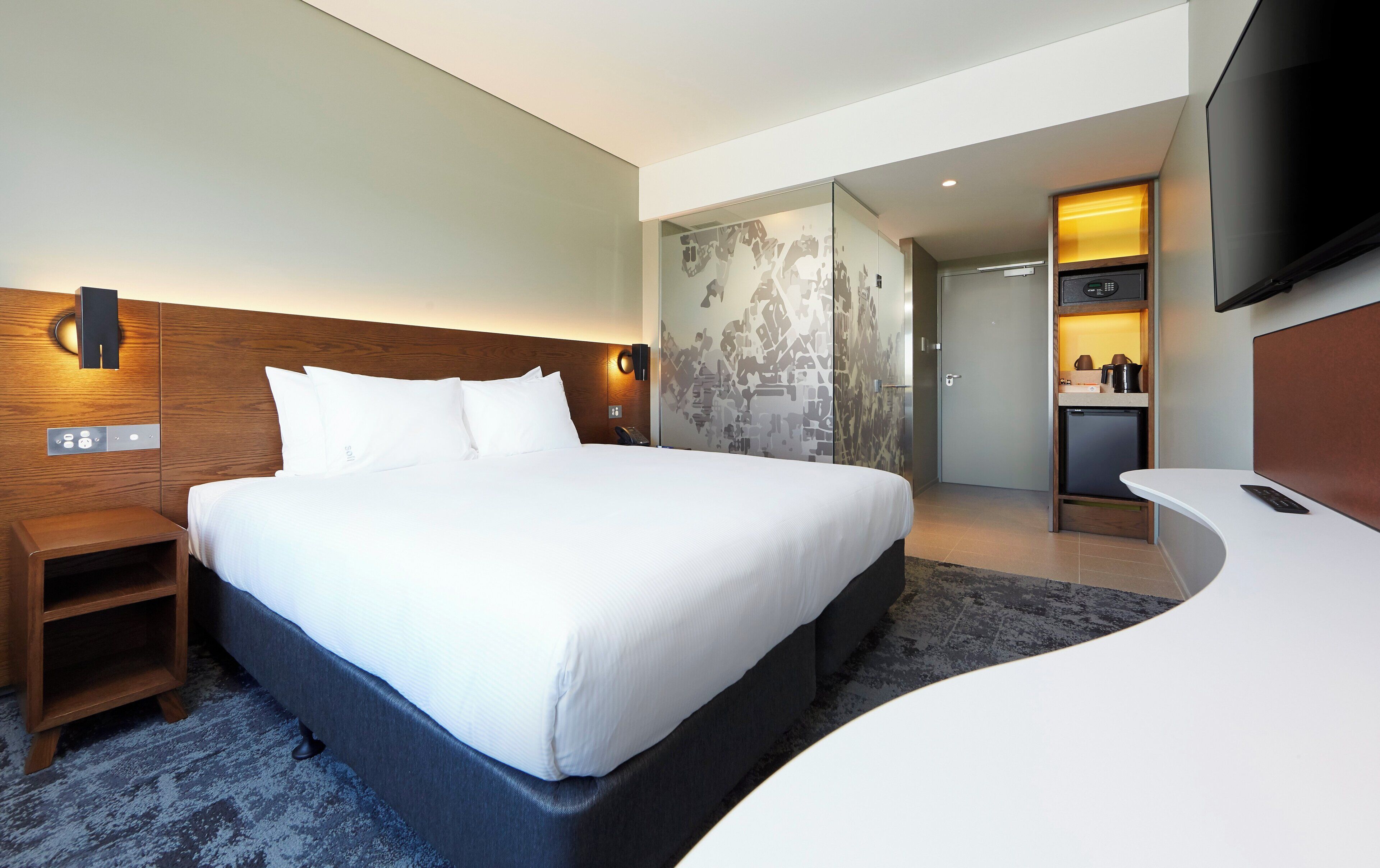 Une chambre d’hôtel moderne dotée d’un grand lit, d’une tête de lit en bois, d’une table de chevet, d’un téléviseur à écran plat fixé au mur et d’un petit coin cuisine.