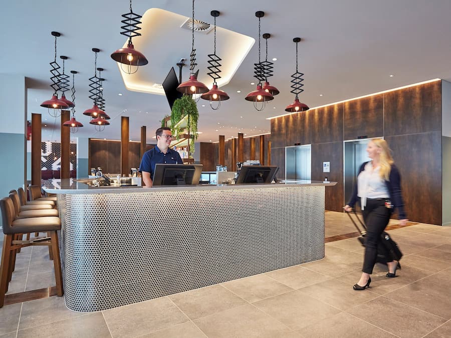 Zona d'entrada d'un hotel amb un disseny modern, que inclou una recepció, un espai d'estar i una gran planta en test.