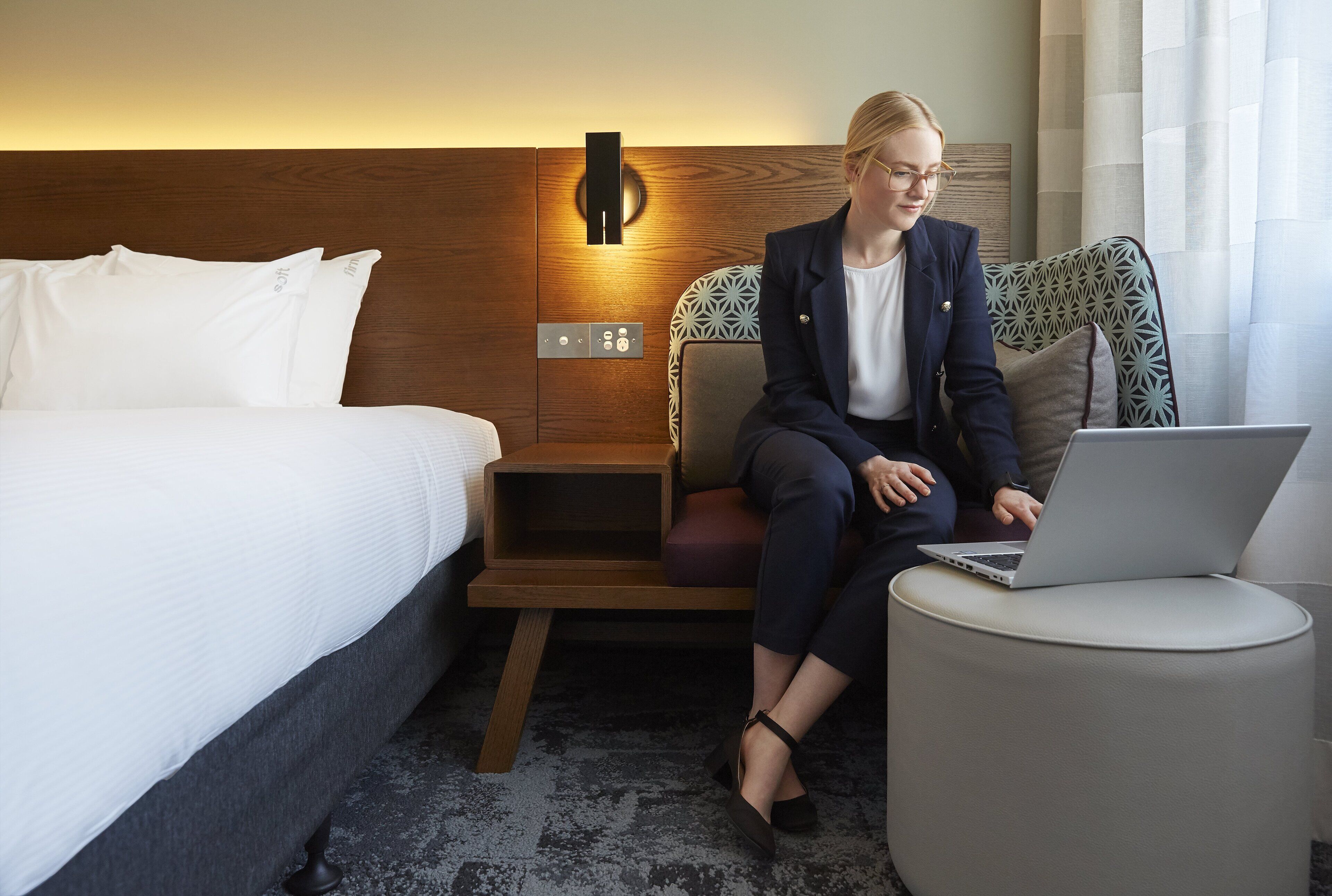 A hotel room with a bed, a desk with a laptop, a nightstand, and a wall-mounted light fixture.
